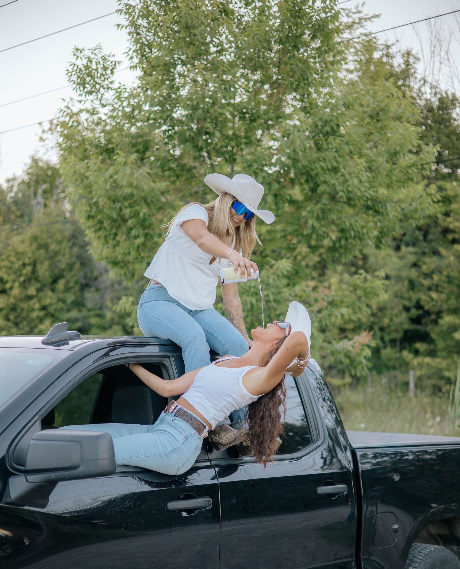 Two women in cowboy hats posing on a black truck outdoors near trees.
