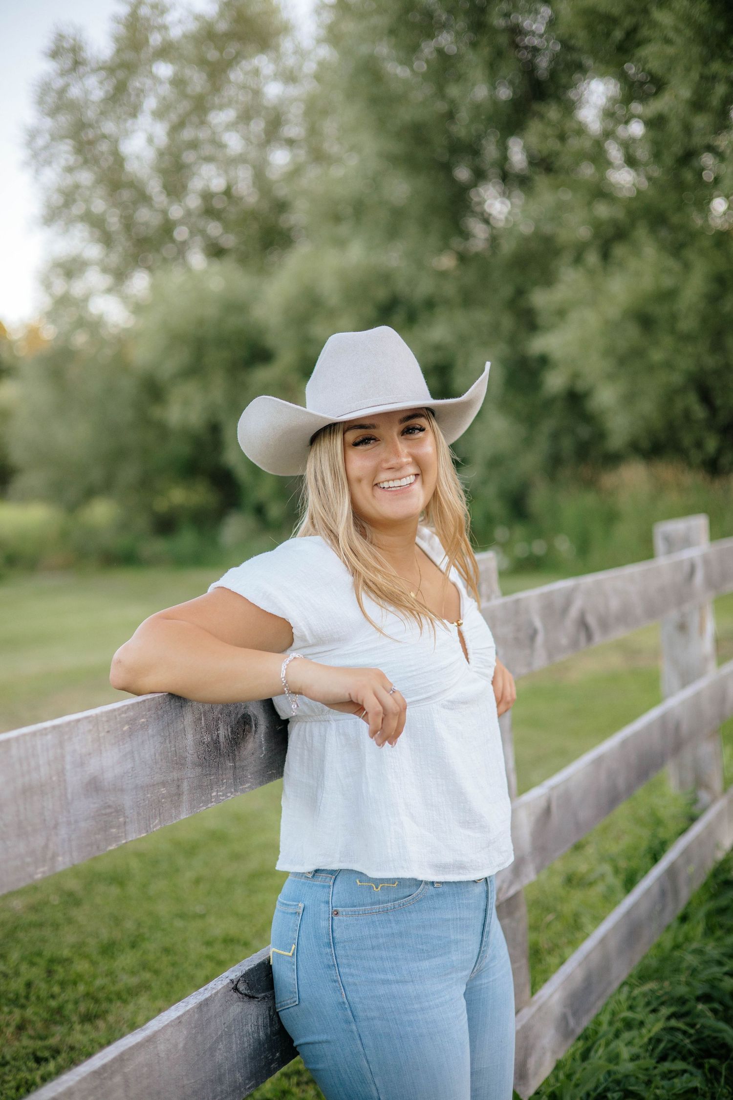 Smiling blonde woman in white top and cowboy hat leaning on a wooden fence.