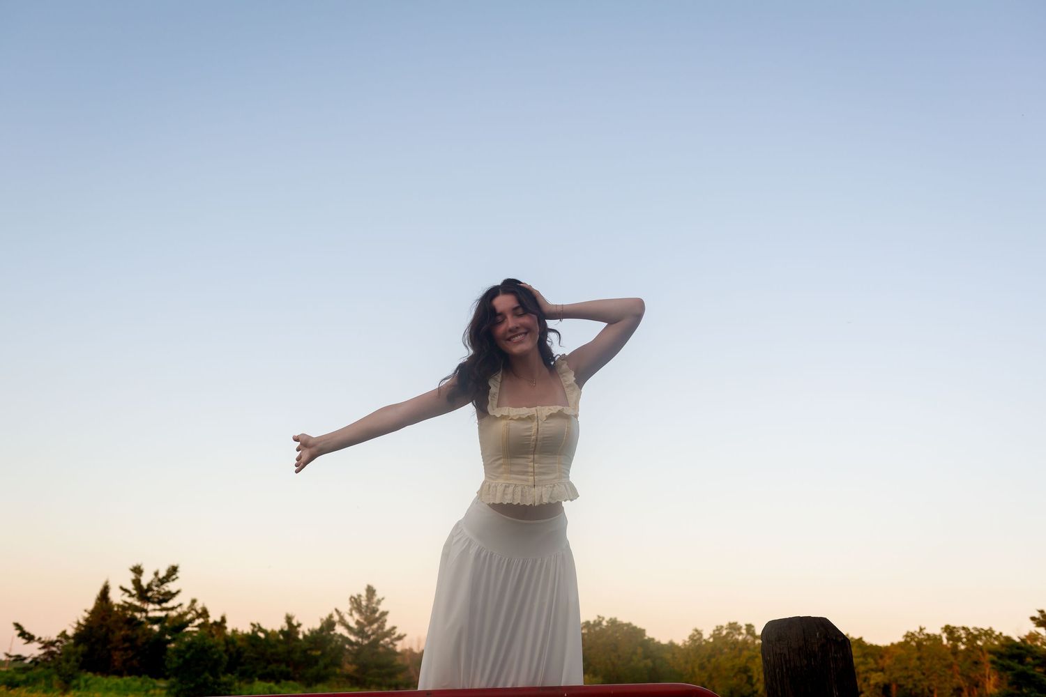 Young woman in white dress posing outdoors at sunset with arms outstretched, smiling joyfully against a clear sky.