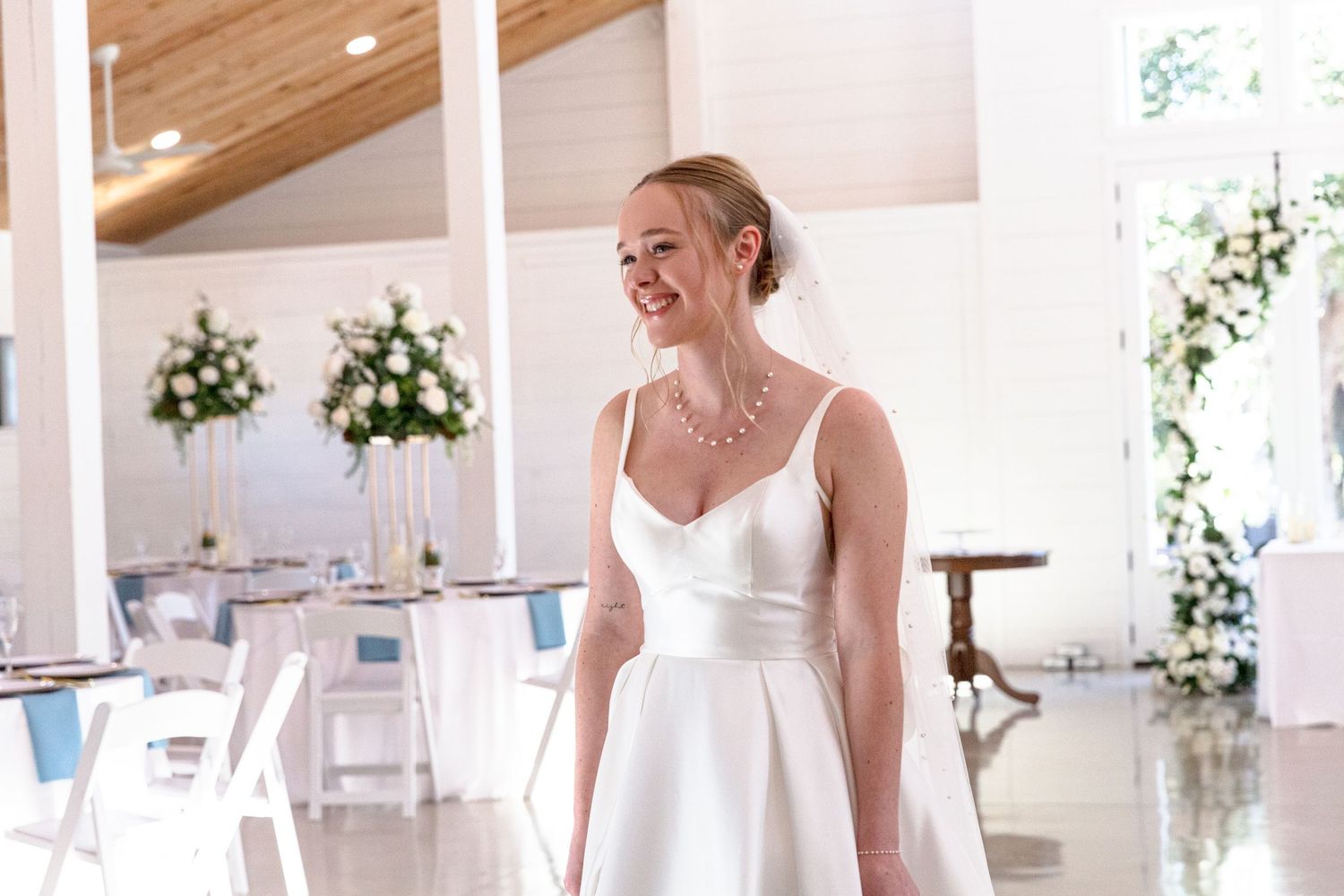 Radiant bride in sleek white gown smiling inside elegant white wedding reception venue.