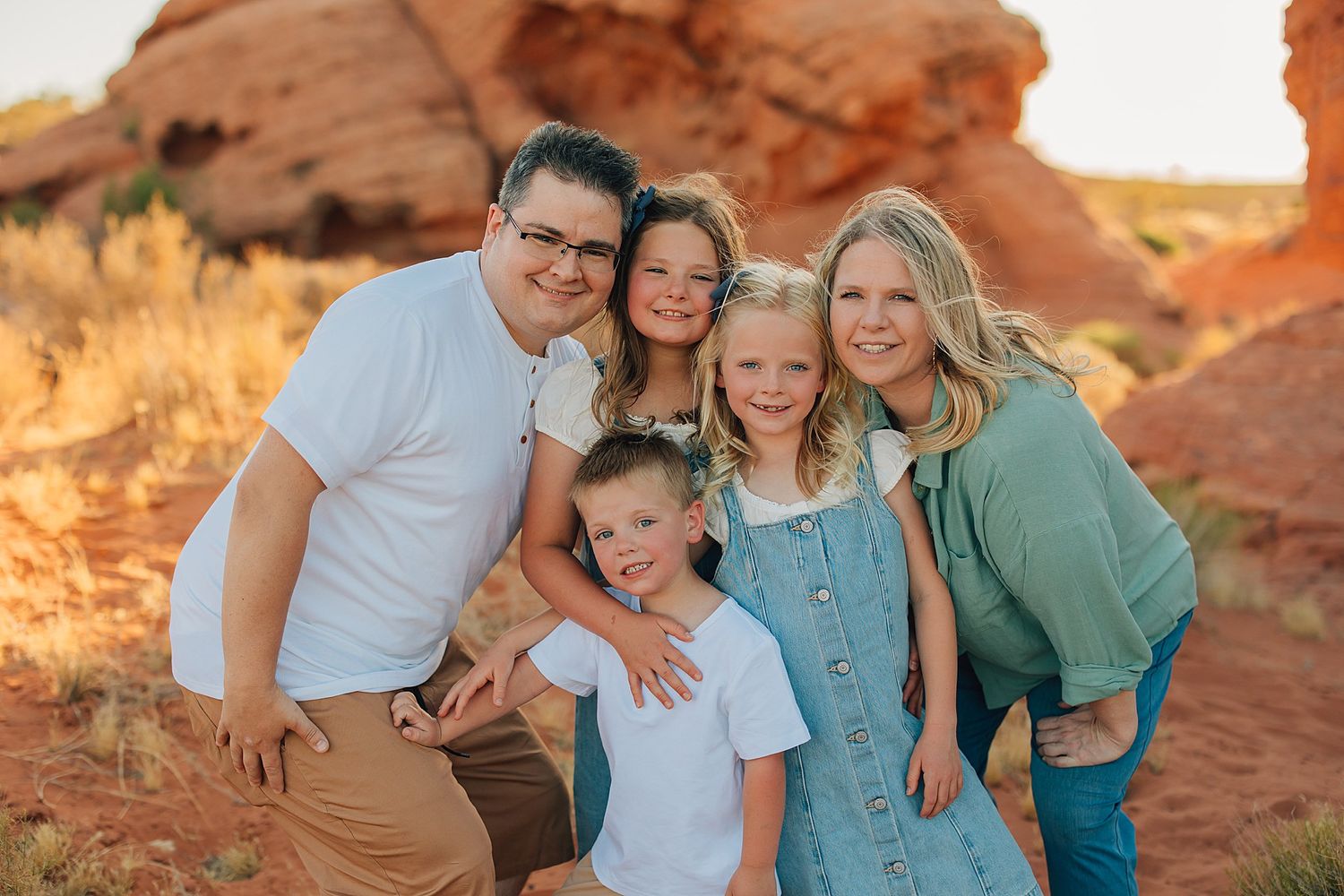 Family of five smiling together in red rock desert landscape during golden hour photography session.