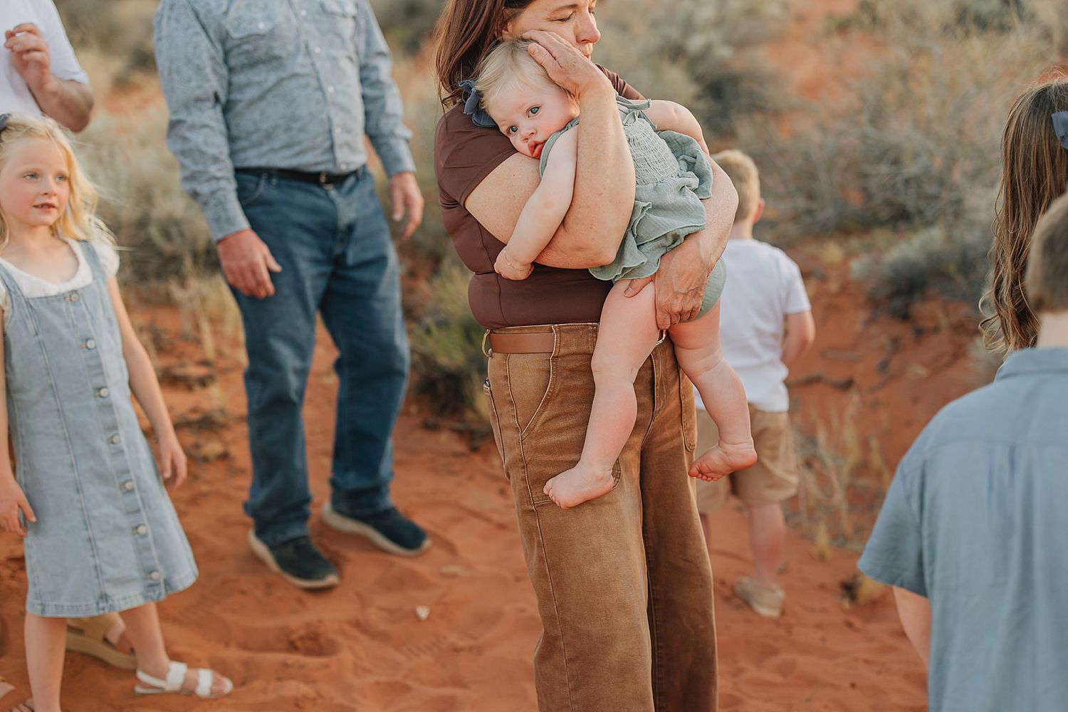 A mother holds her baby outdoors on red desert sand surrounded by family during a professional photo session.