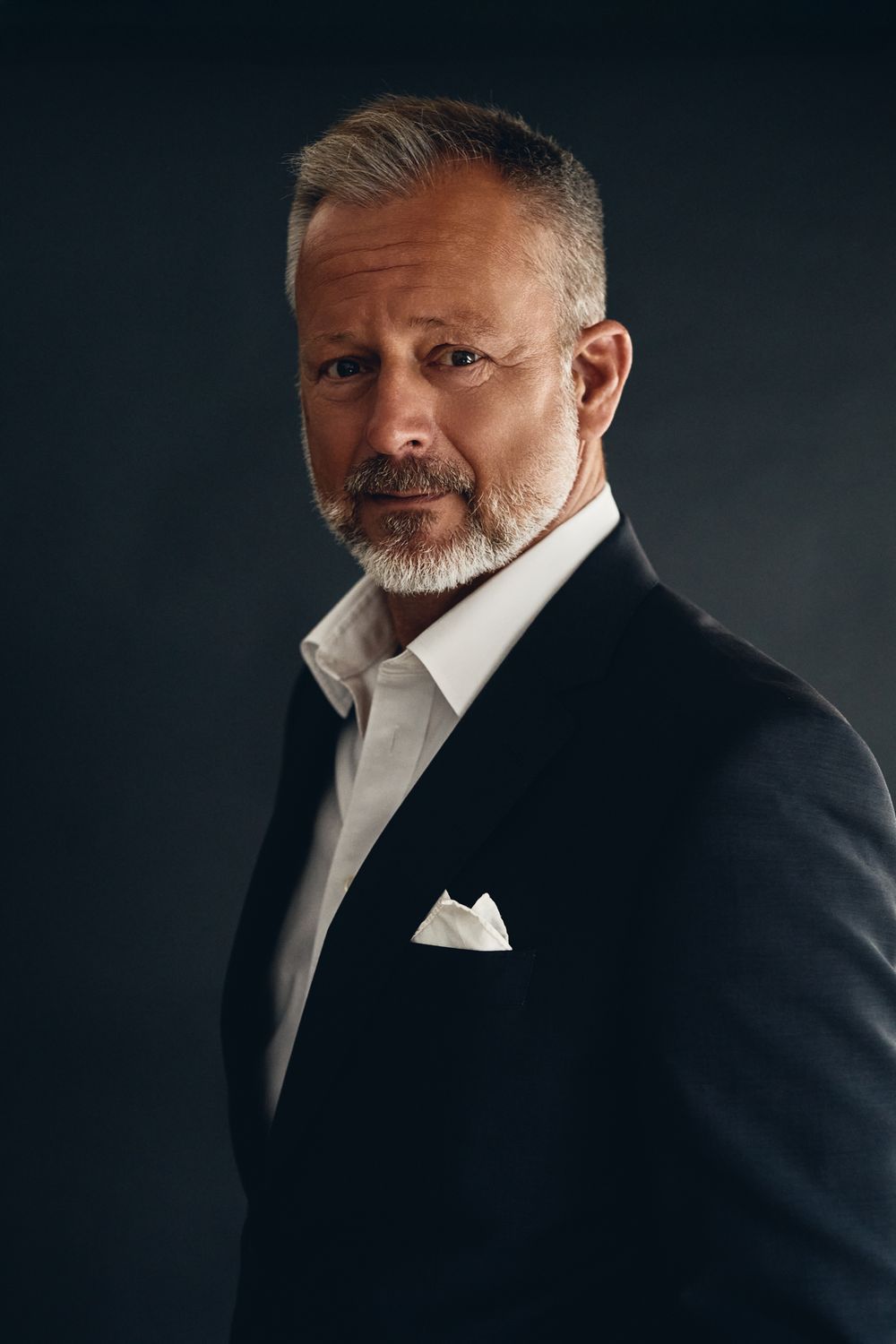 Distinguished older man with grey hair and beard in a dark suit with white pocket square on dark background.