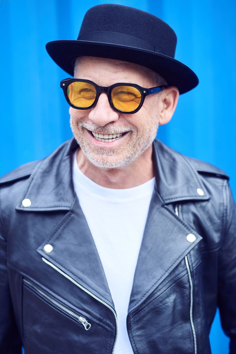 Cheerful older man wearing yellow sunglasses, black hat, and leather jacket smiling against blue background.