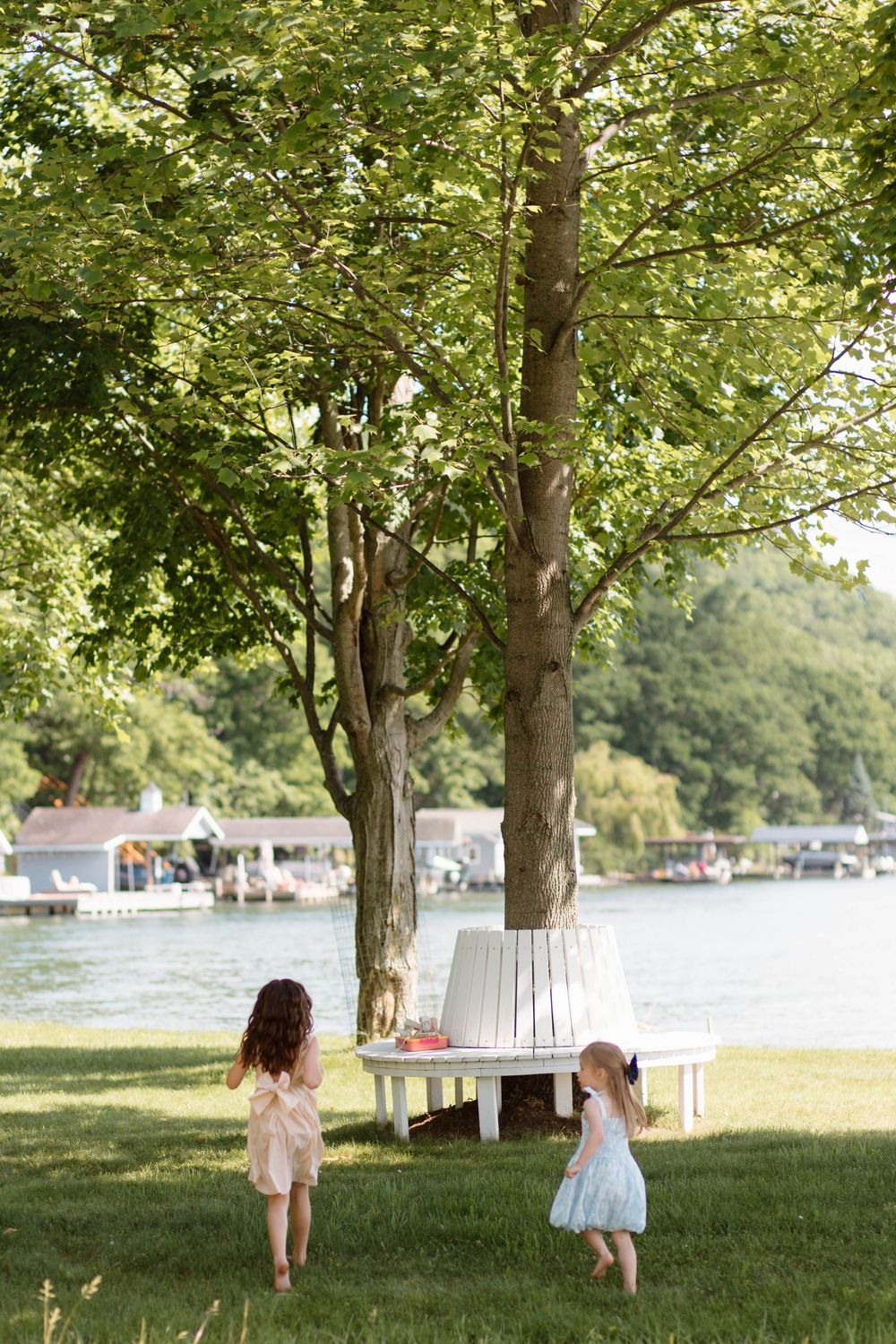 Two young girls in summer dresses play near a white tree bench by a lakeside park on a sunny day.