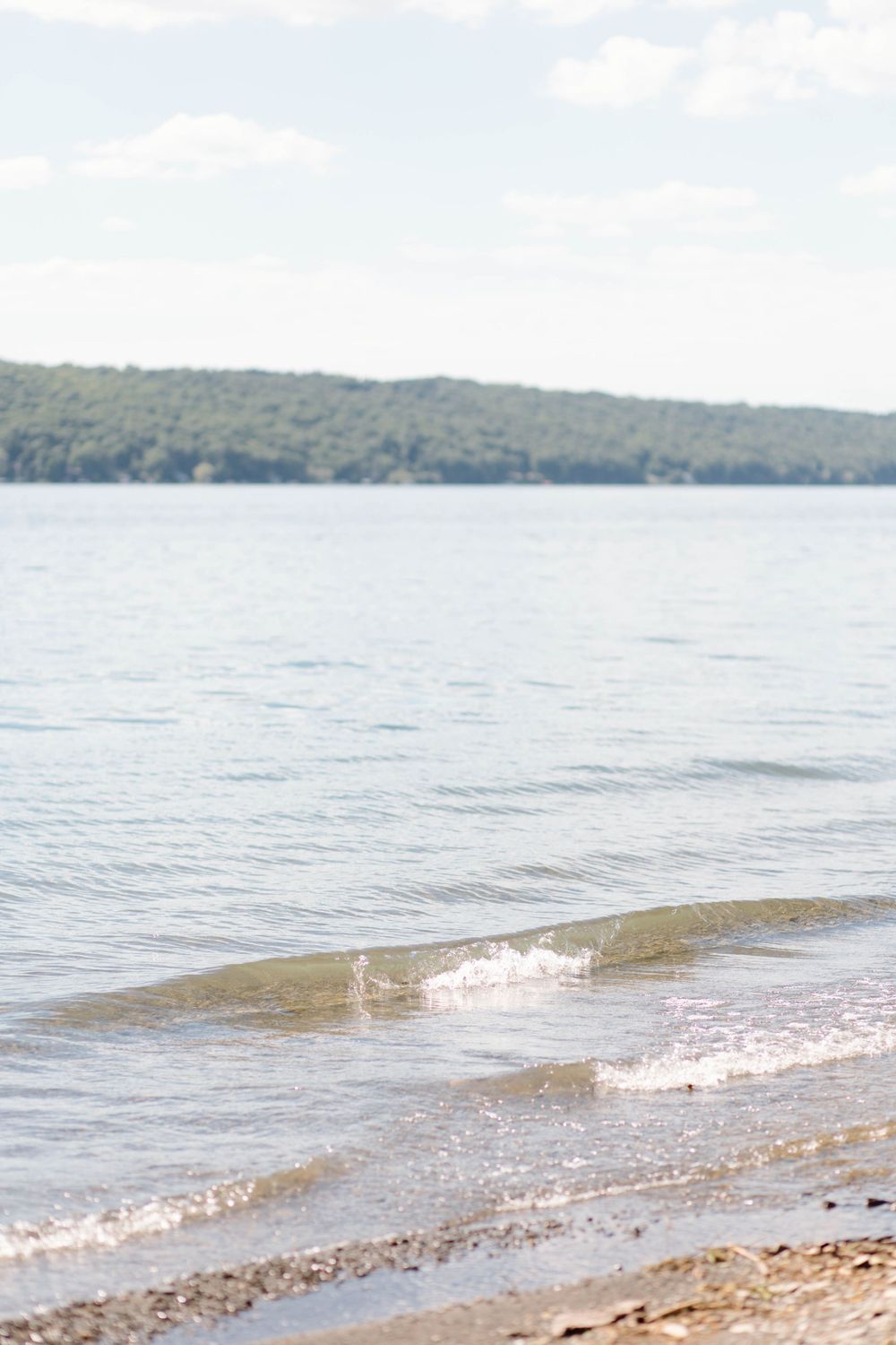 Calm lake shoreline with gentle waves, sandy beach, and tree-covered hills in the distance.