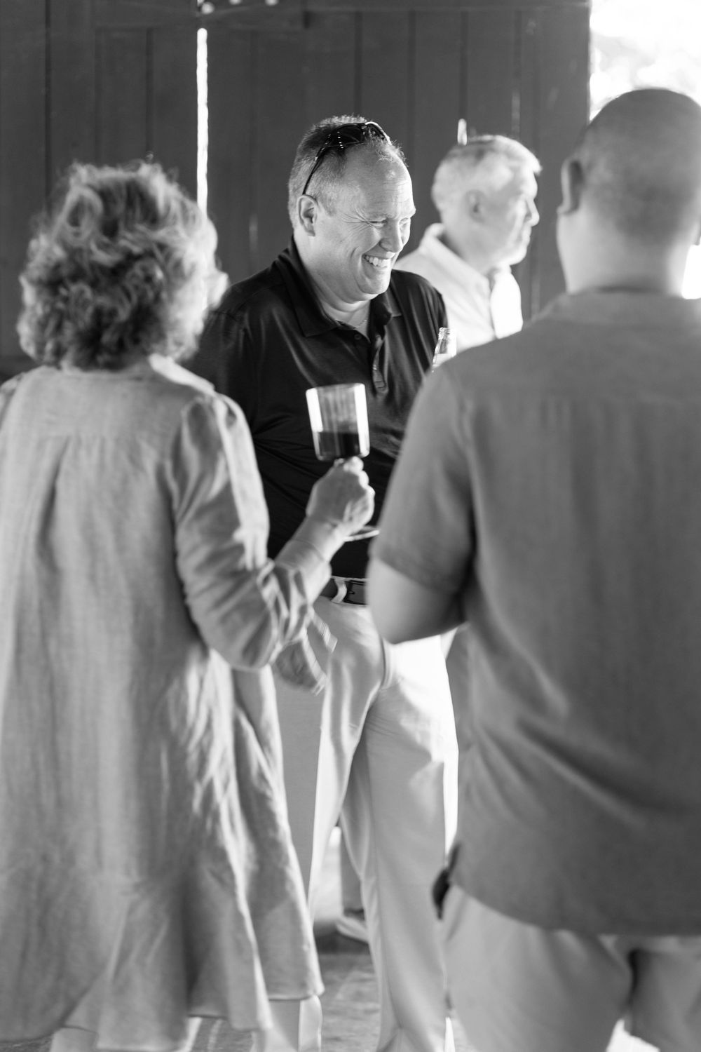 Black and white photo of smiling man holding wine glass socializing with guests at an outdoor gathering.