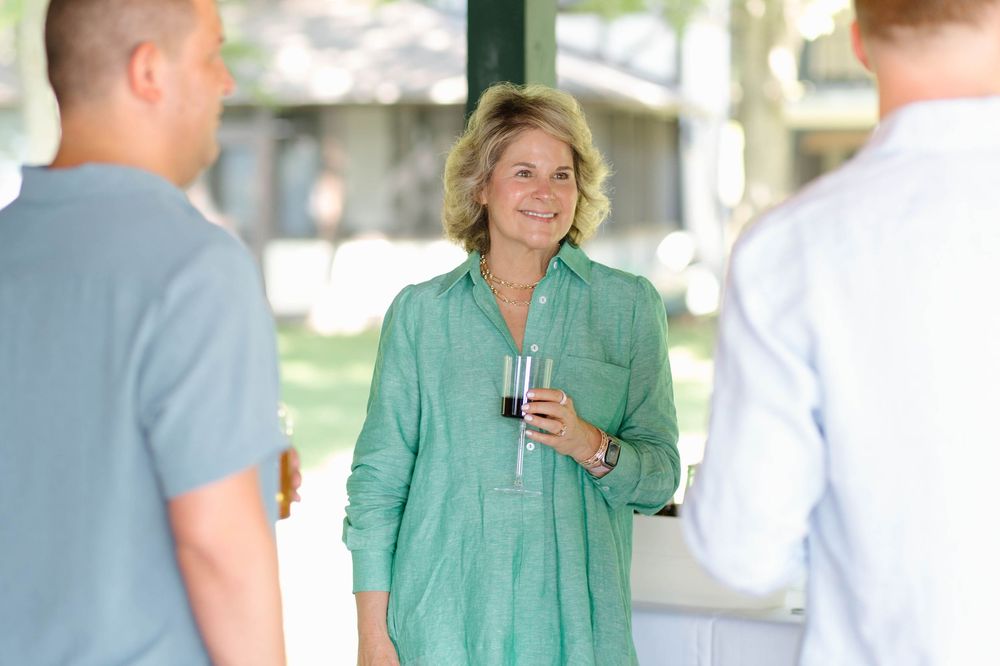Woman in green shirt smiling and holding red wine glass at an outdoor social gathering.