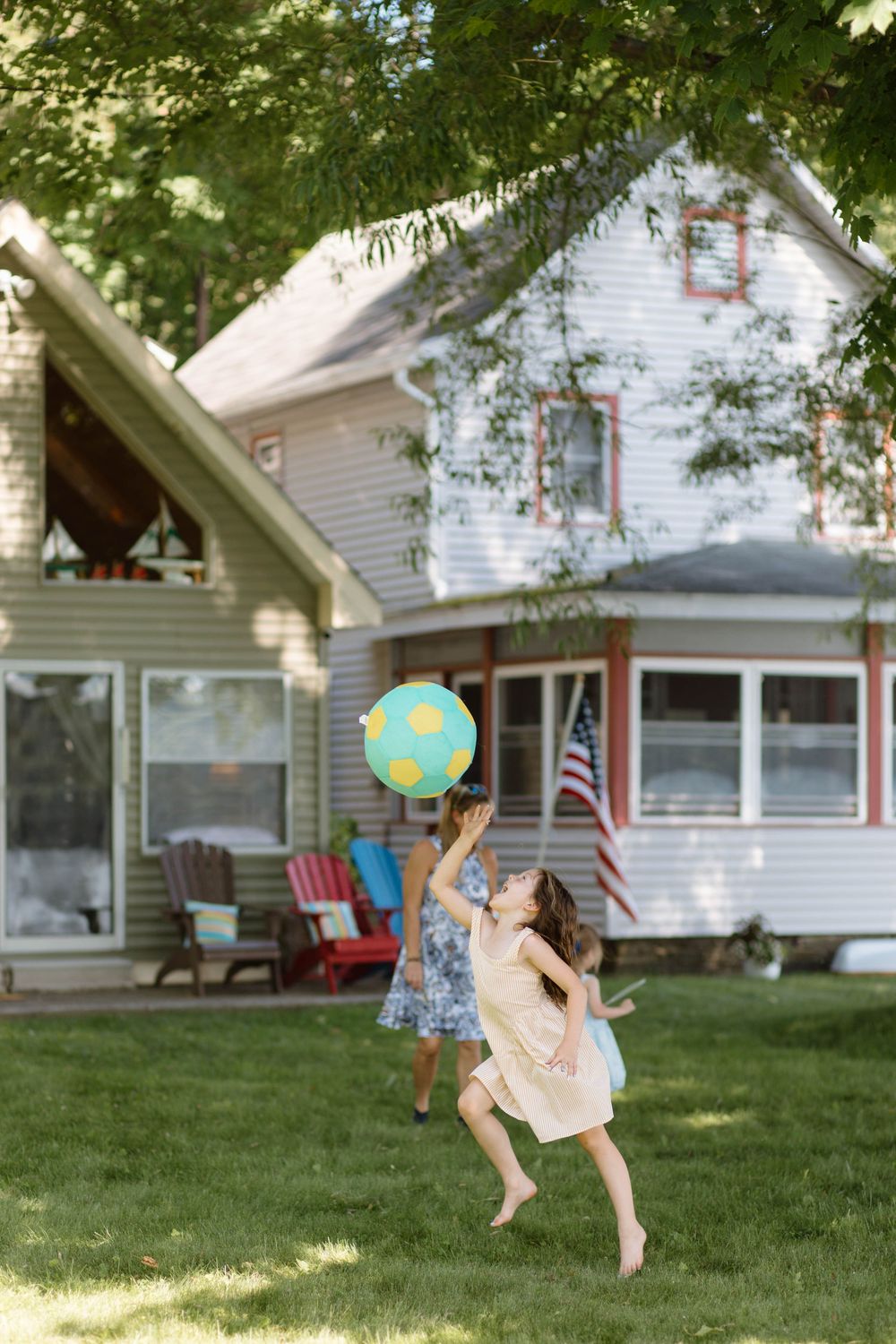 Two girls playing with a colorful beach ball in a green backyard beside a white American farmhouse.