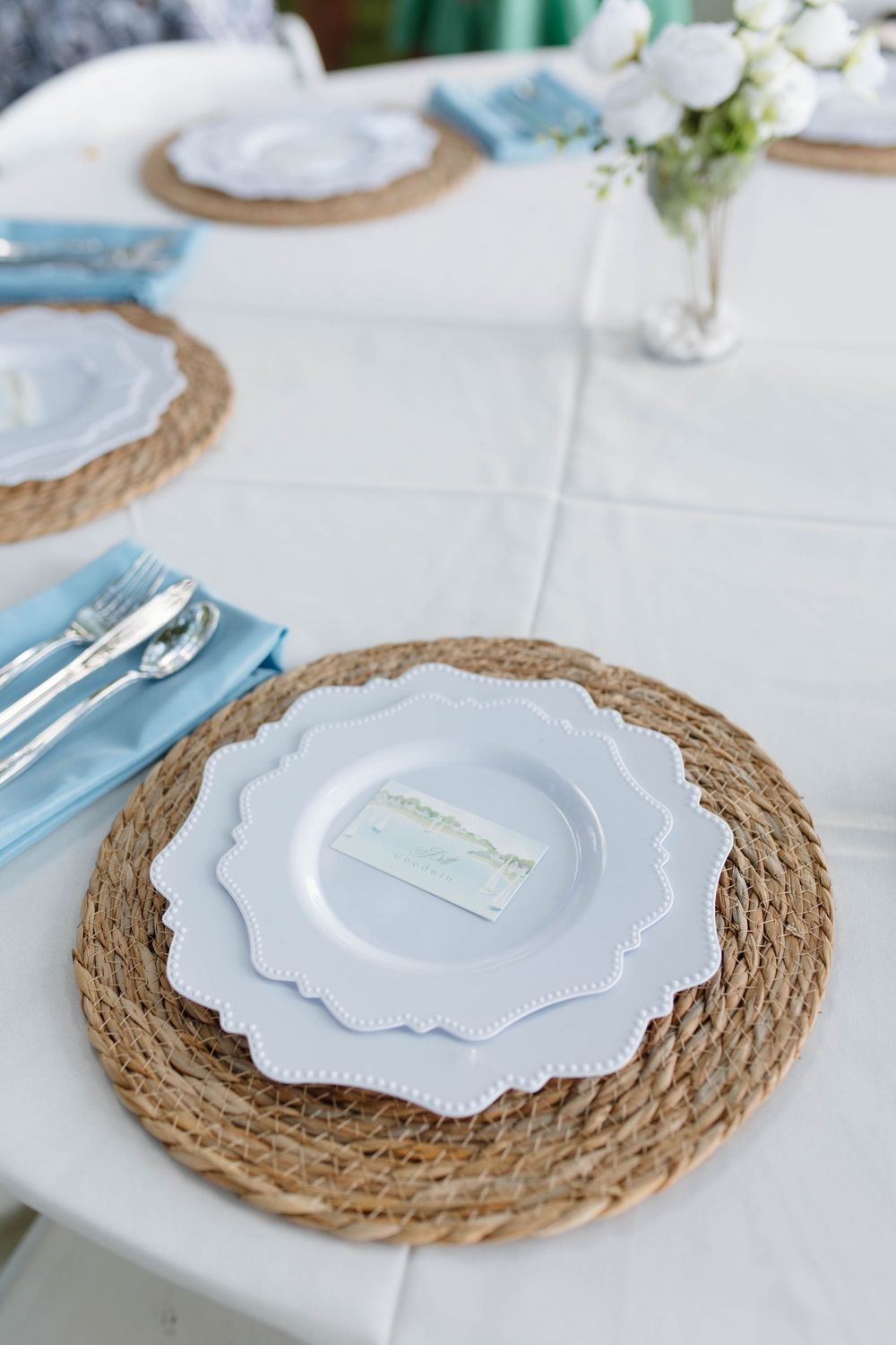Elegant table setting with woven placemats, white scalloped plates, blue napkins, and silver cutlery.