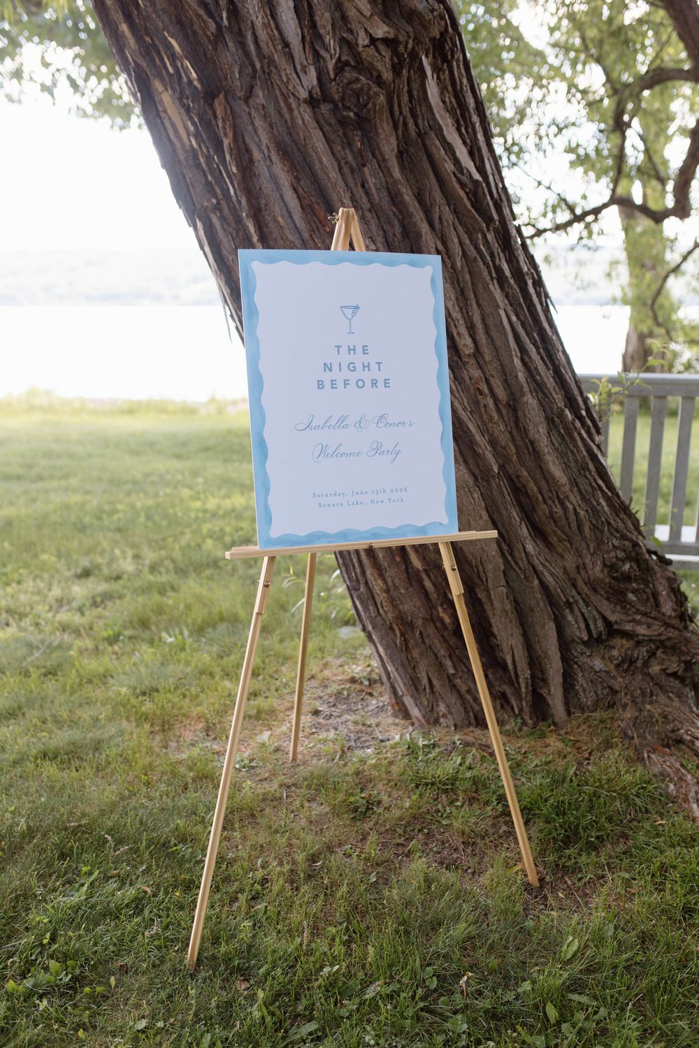 Elegant wedding welcome sign on wooden easel reading The Night Before rehearsal party leaning against tree outdoors.