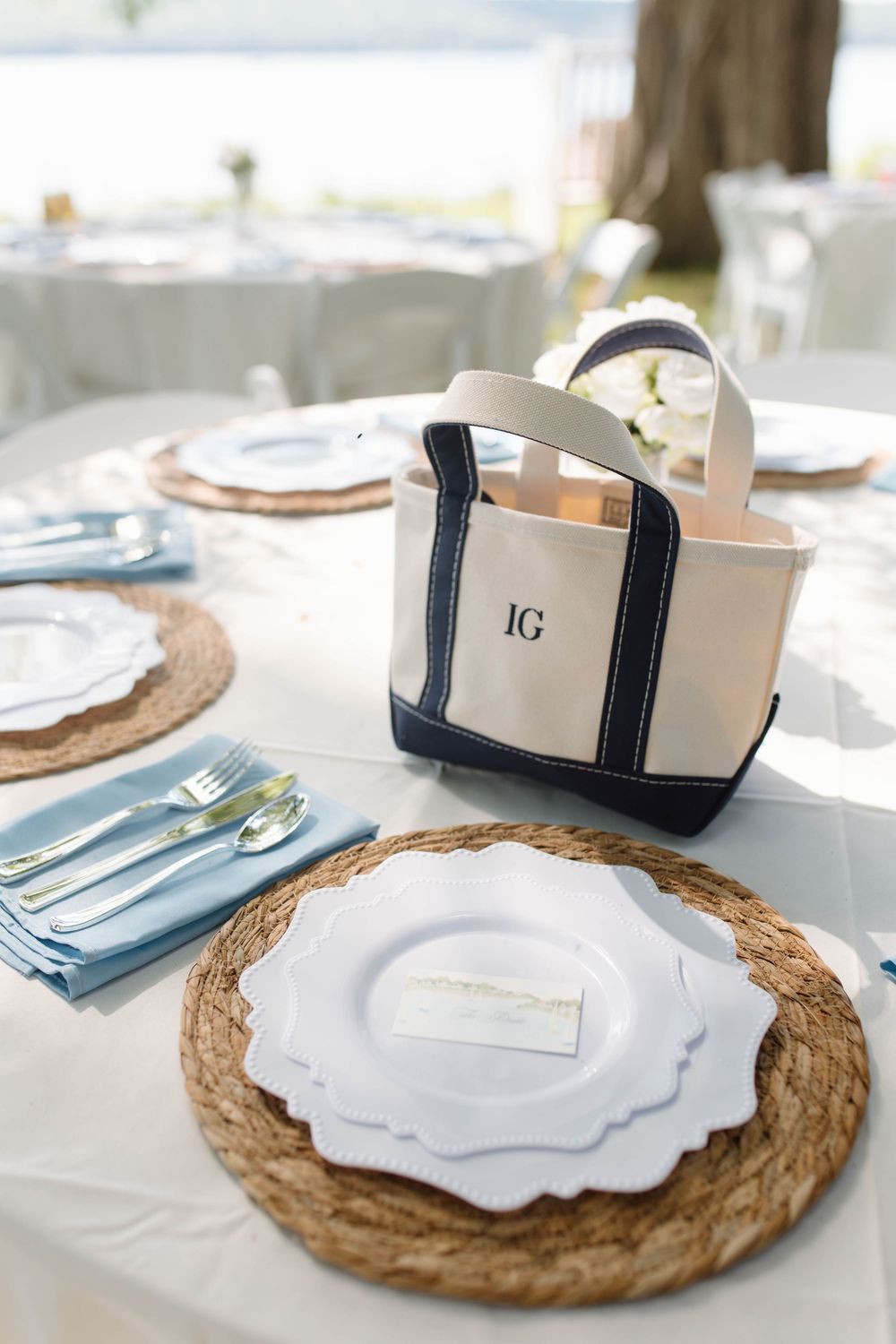 Elegant outdoor wedding table setting with woven charger plates, blue napkins, gold silverware, and a monogrammed tote bag favor.