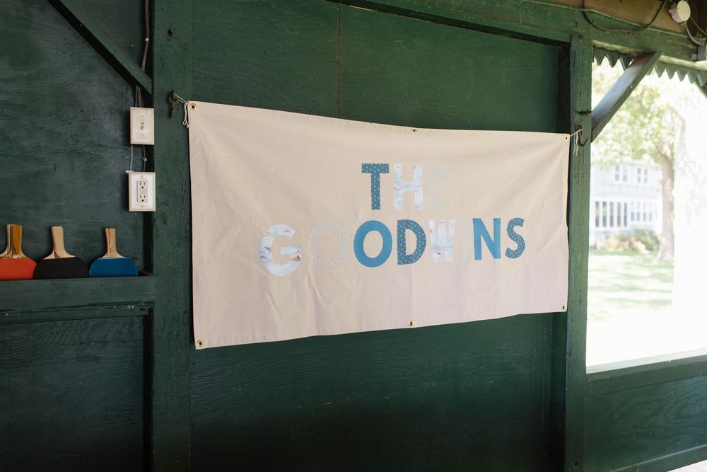 A white banner reading 'The Goodins' hangs on a green wooden wall at an outdoor event.
