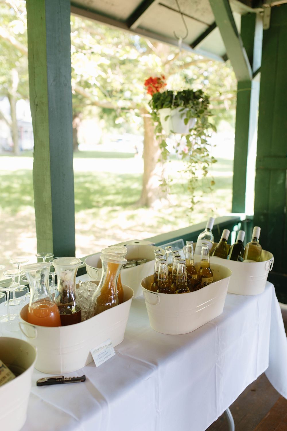 Outdoor beverage station with white buckets filled with wine bottles and juice pitchers on a white-draped table under a green pergola.