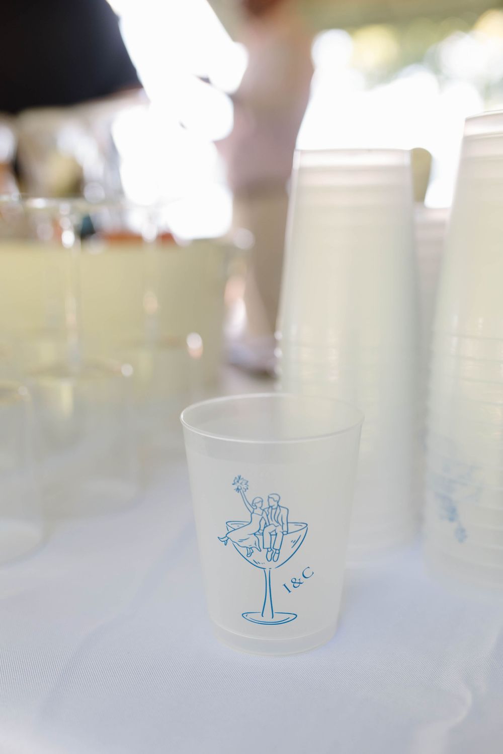 Frosted plastic cup with blue martini glass illustration and initials I&C on a white table at an outdoor wedding reception.
