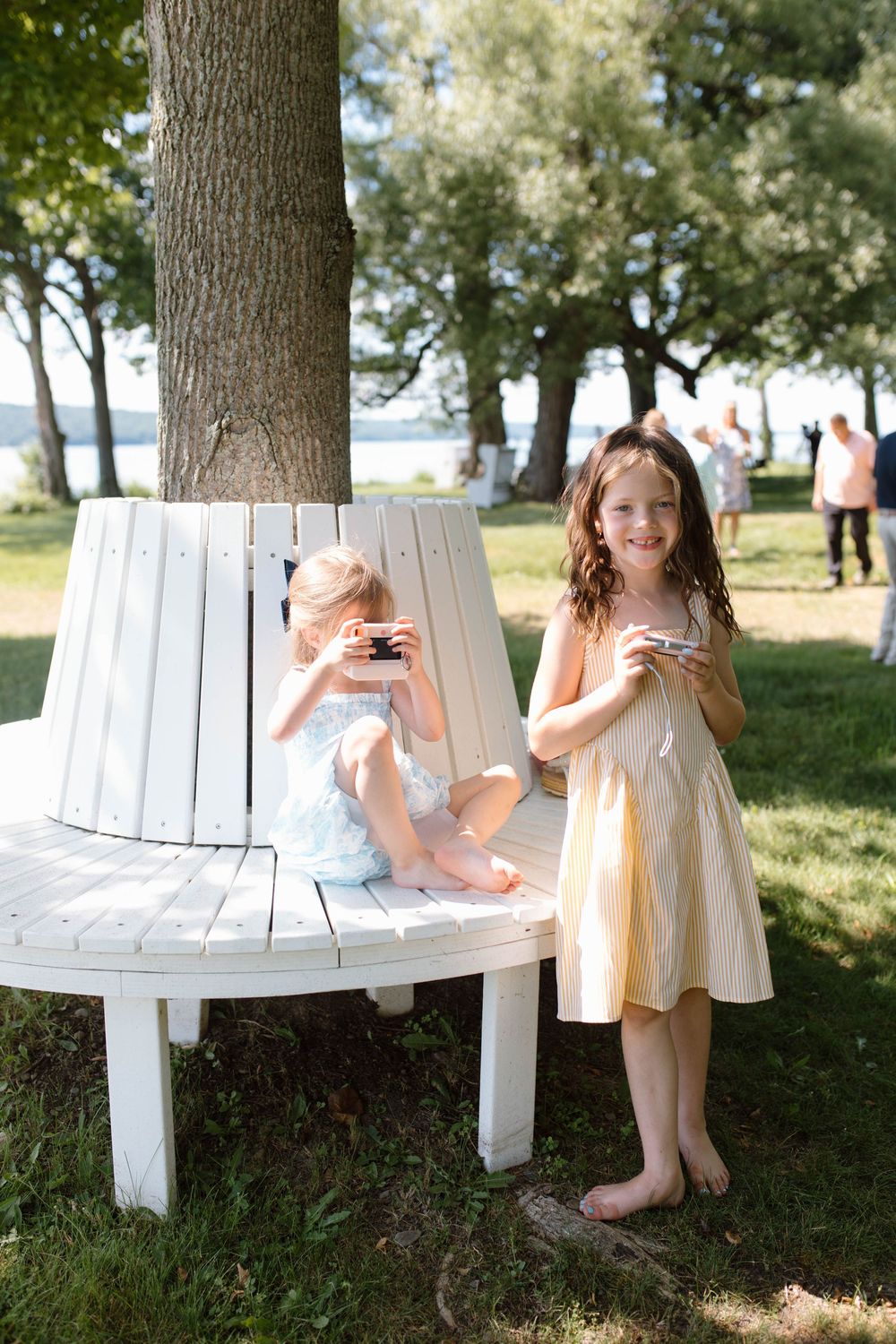Two young girls in summer dresses eating cake near a white tree bench at an outdoor event.