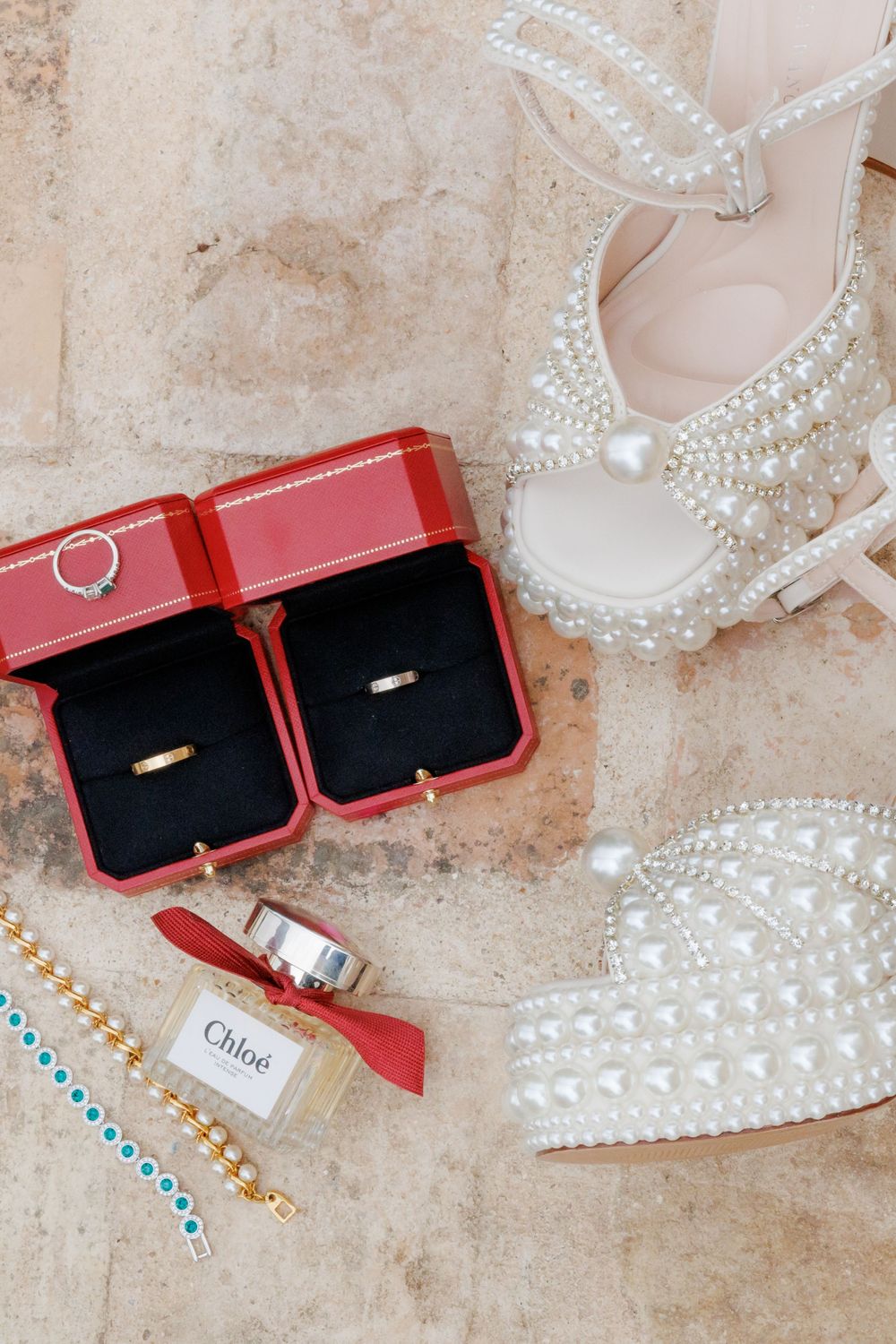 Pearl-embellished bridal heels beside Cartier wedding rings in red boxes, Chloe perfume, and turquoise bracelet on marble.