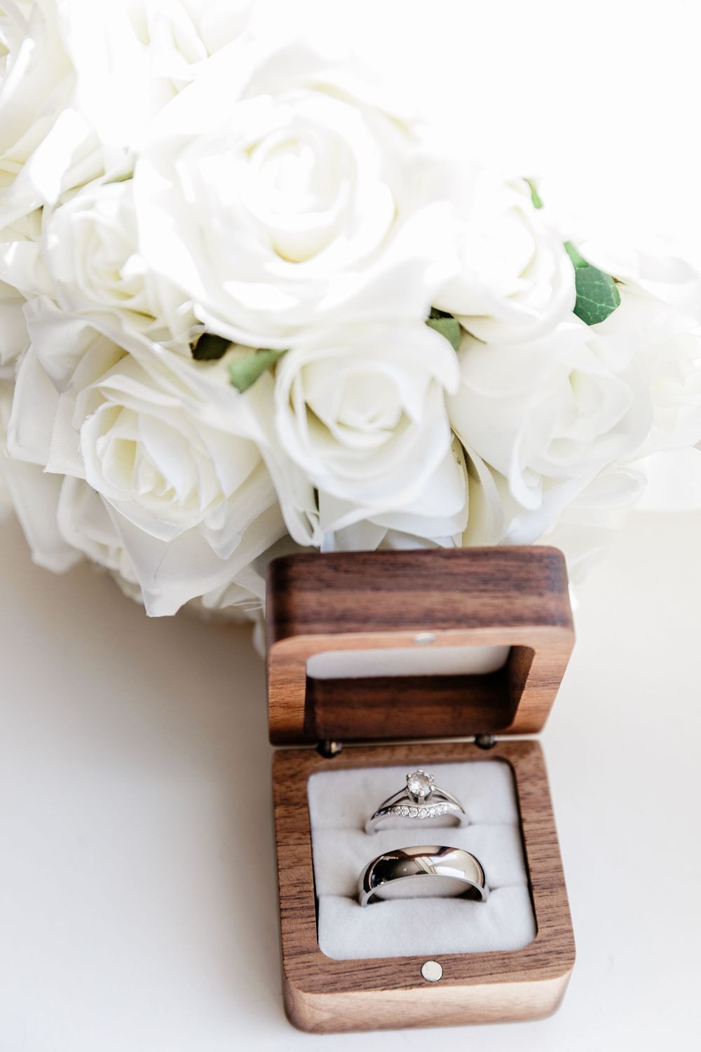 Elegant wedding rings in a wooden box beside a white rose bouquet on a white surface.
