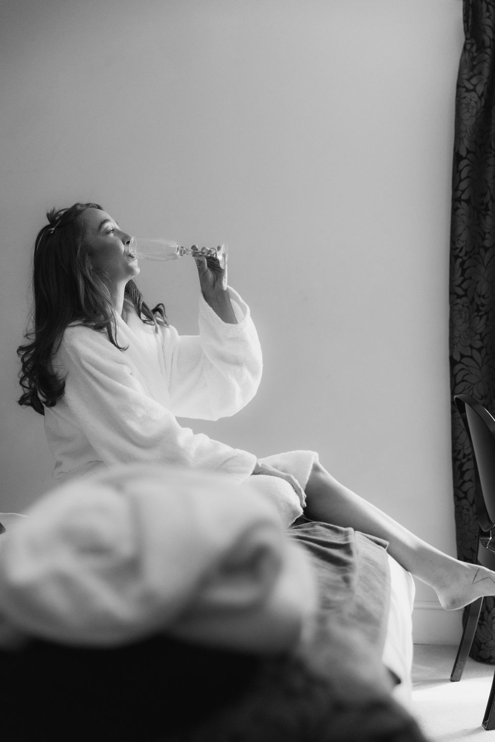 Woman in white bathrobe sitting on bed drinking champagne in black and white photo.