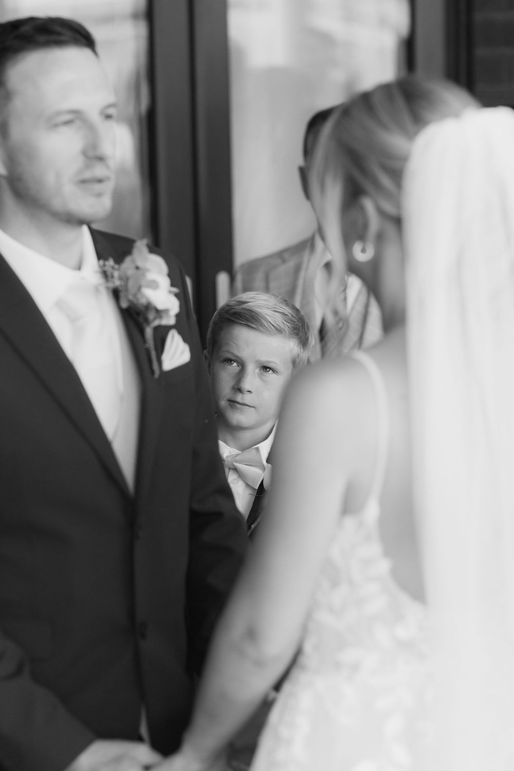 Black and white wedding photo of a young boy in a suit watching the bride and groom exchange vows at the altar.