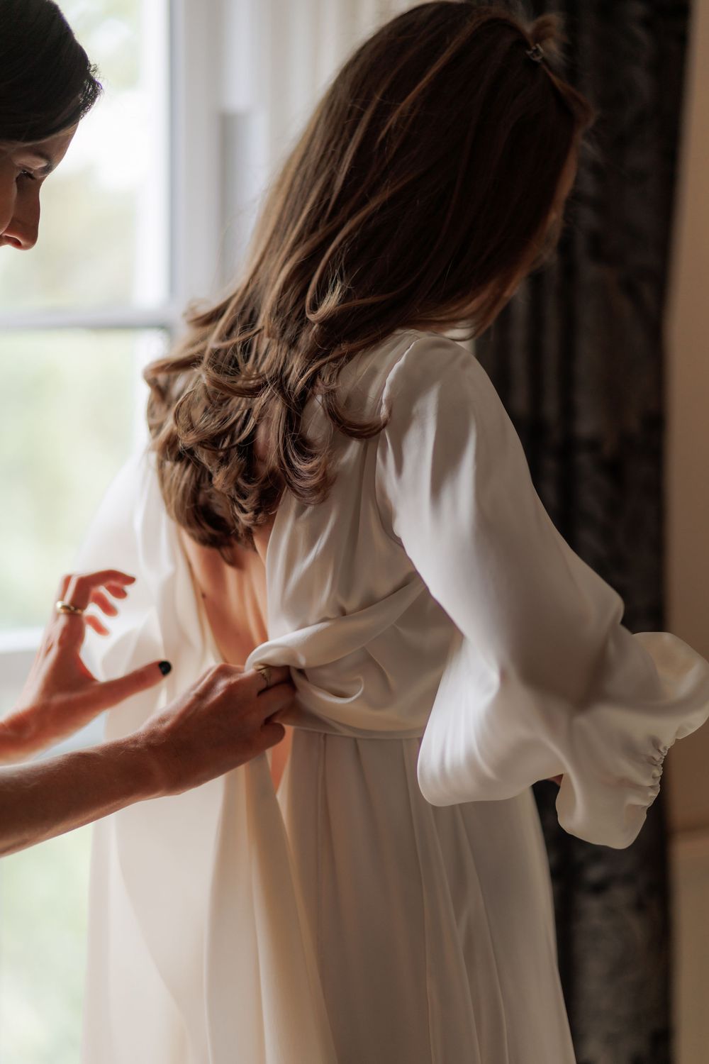 Bride in white long-sleeve gown being helped with dress buttons near a bright window on wedding day.
