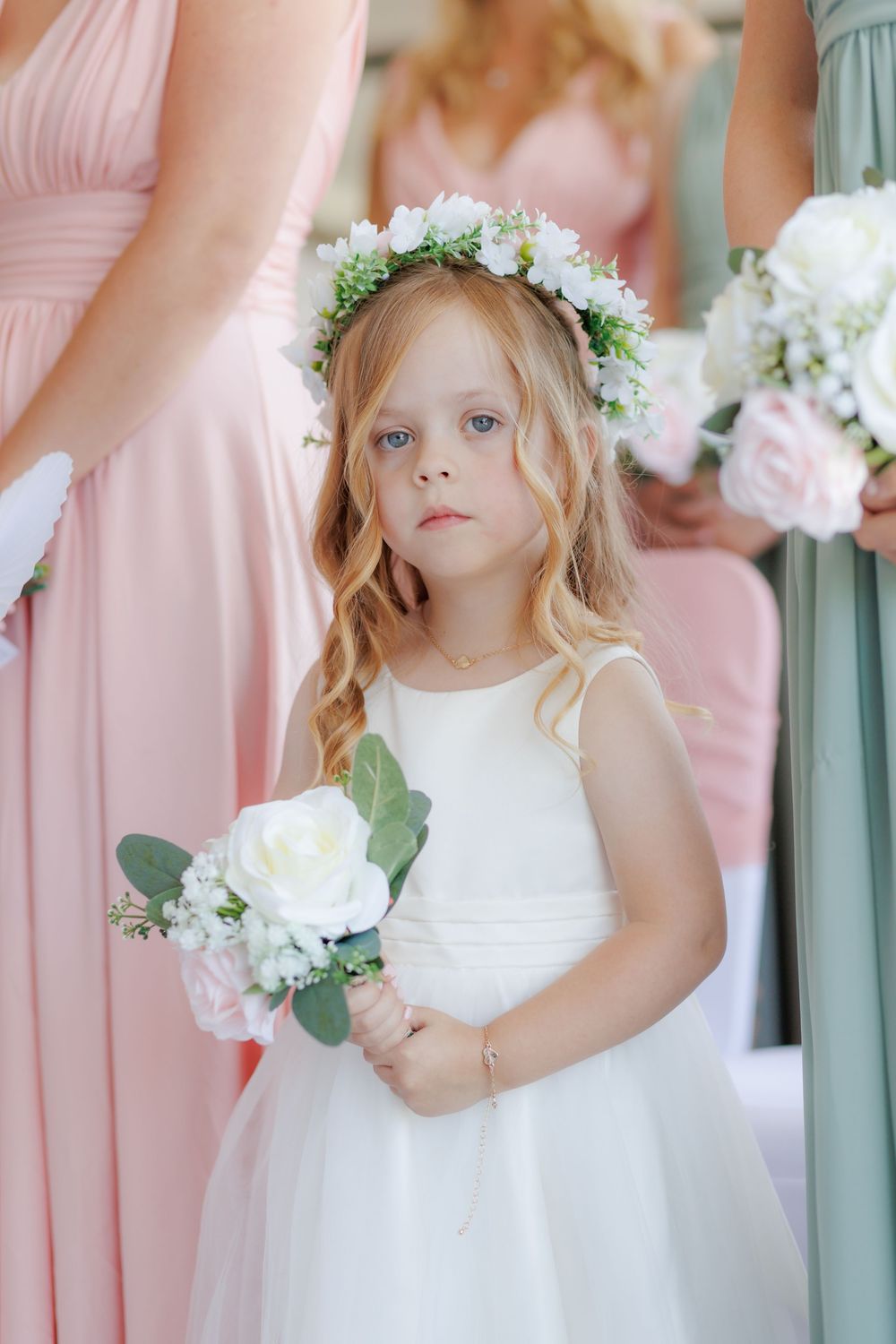 A young flower girl in a white dress and floral crown holds a bouquet, standing beside bridesmaids in pink gowns.