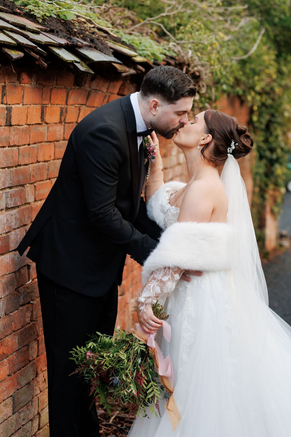 Bride and groom share a romantic kiss beside a rustic brick wall, bride wearing a white gown with fur wrap holding a bouquet.