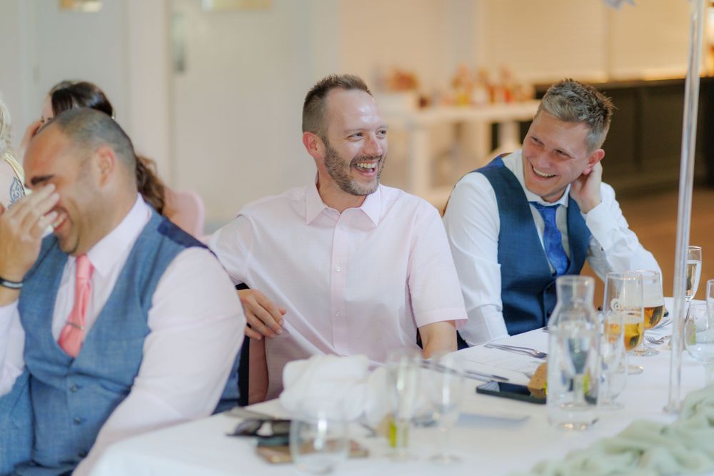 Three men in smart casual attire laughing joyfully at a wedding reception dinner table.