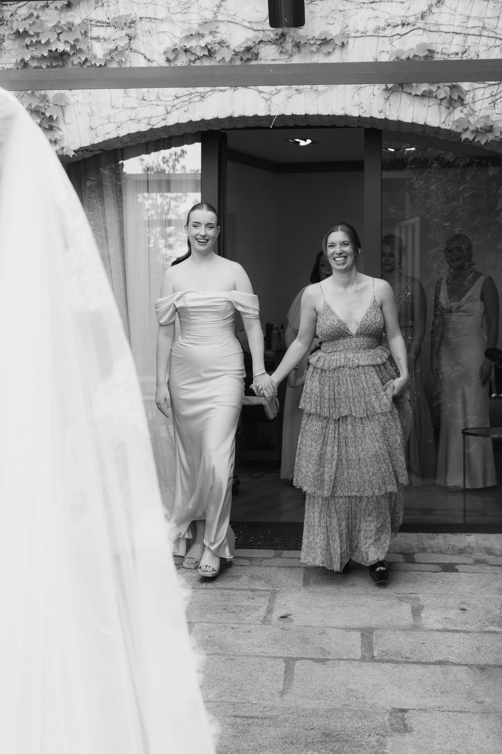 Two women in elegant gowns hold hands smiling as they walk through a stone archway at a wedding venue.