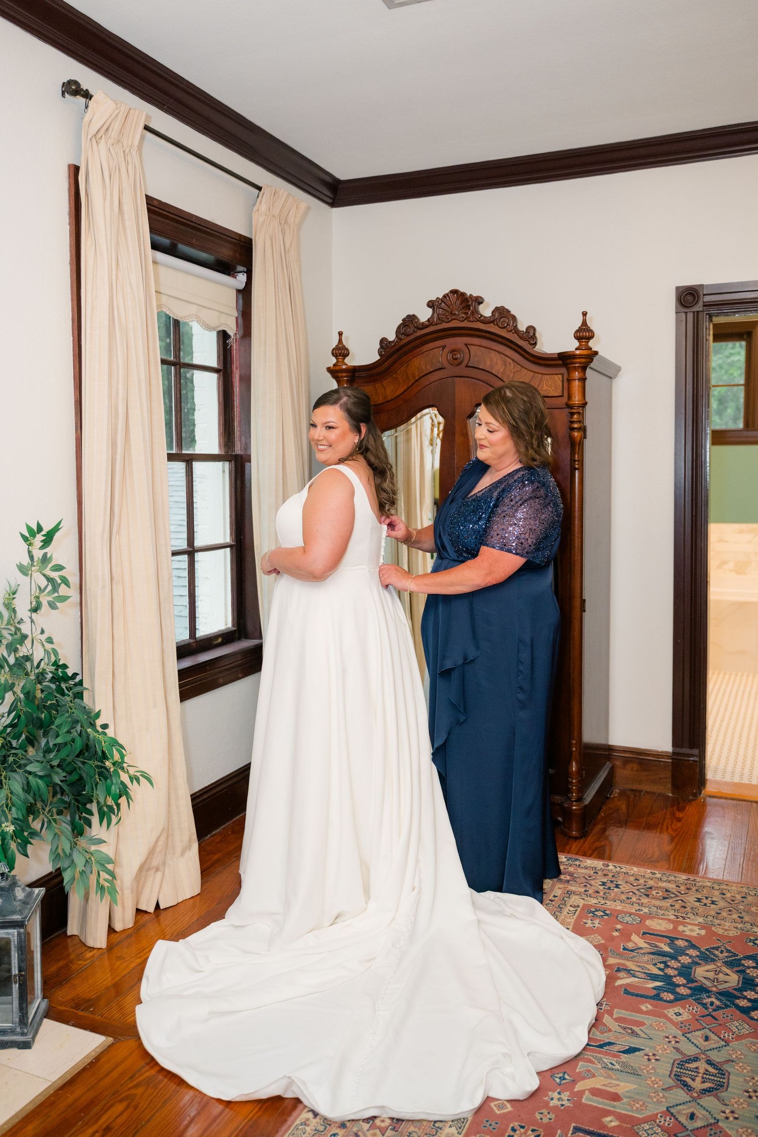 Bride in white gown being helped by woman in navy dress near antique armoire in elegant room.