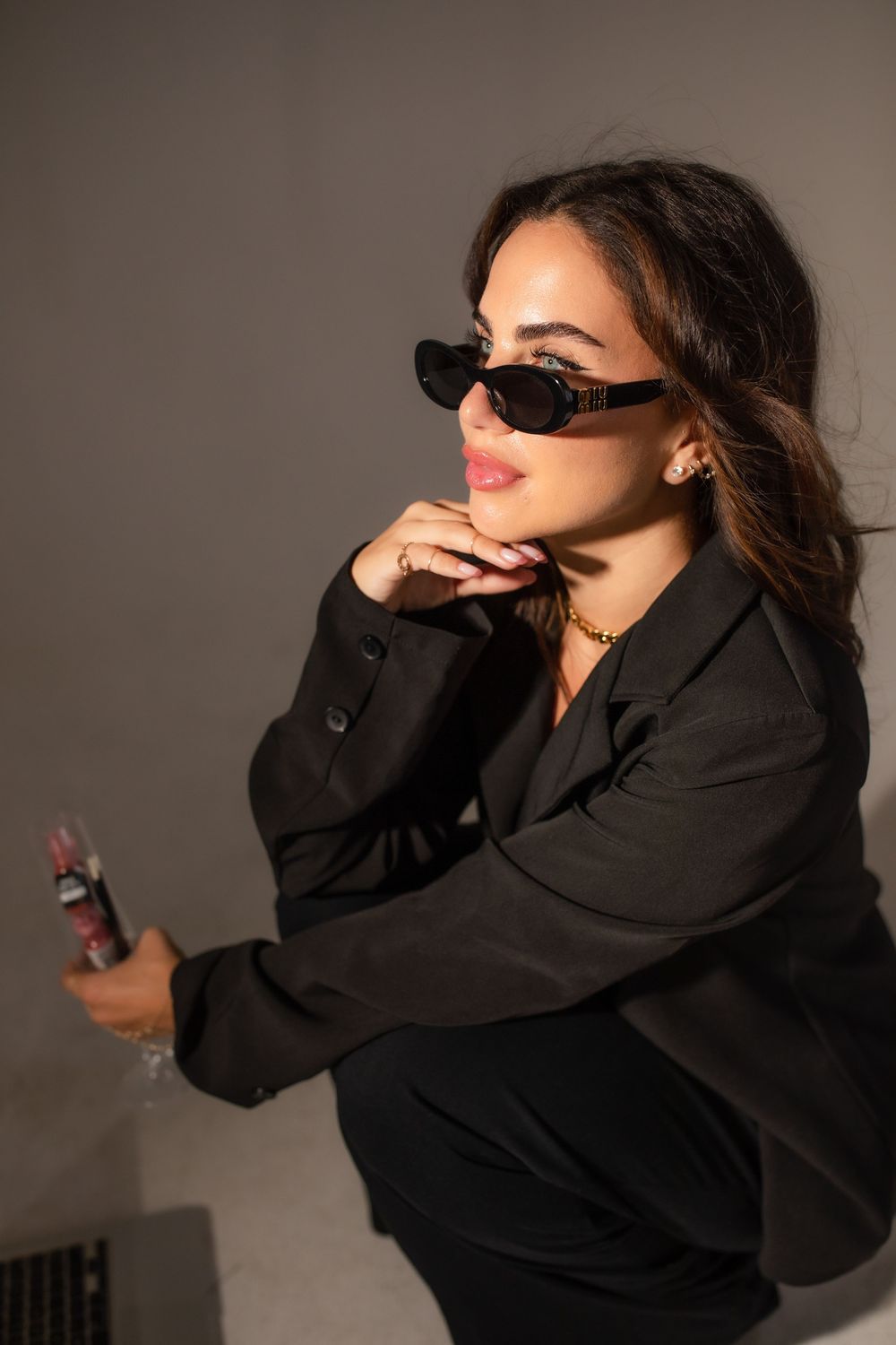 A stylish woman in a black blazer and sunglasses poses confidently, holding a phone, with gold jewelry accents.