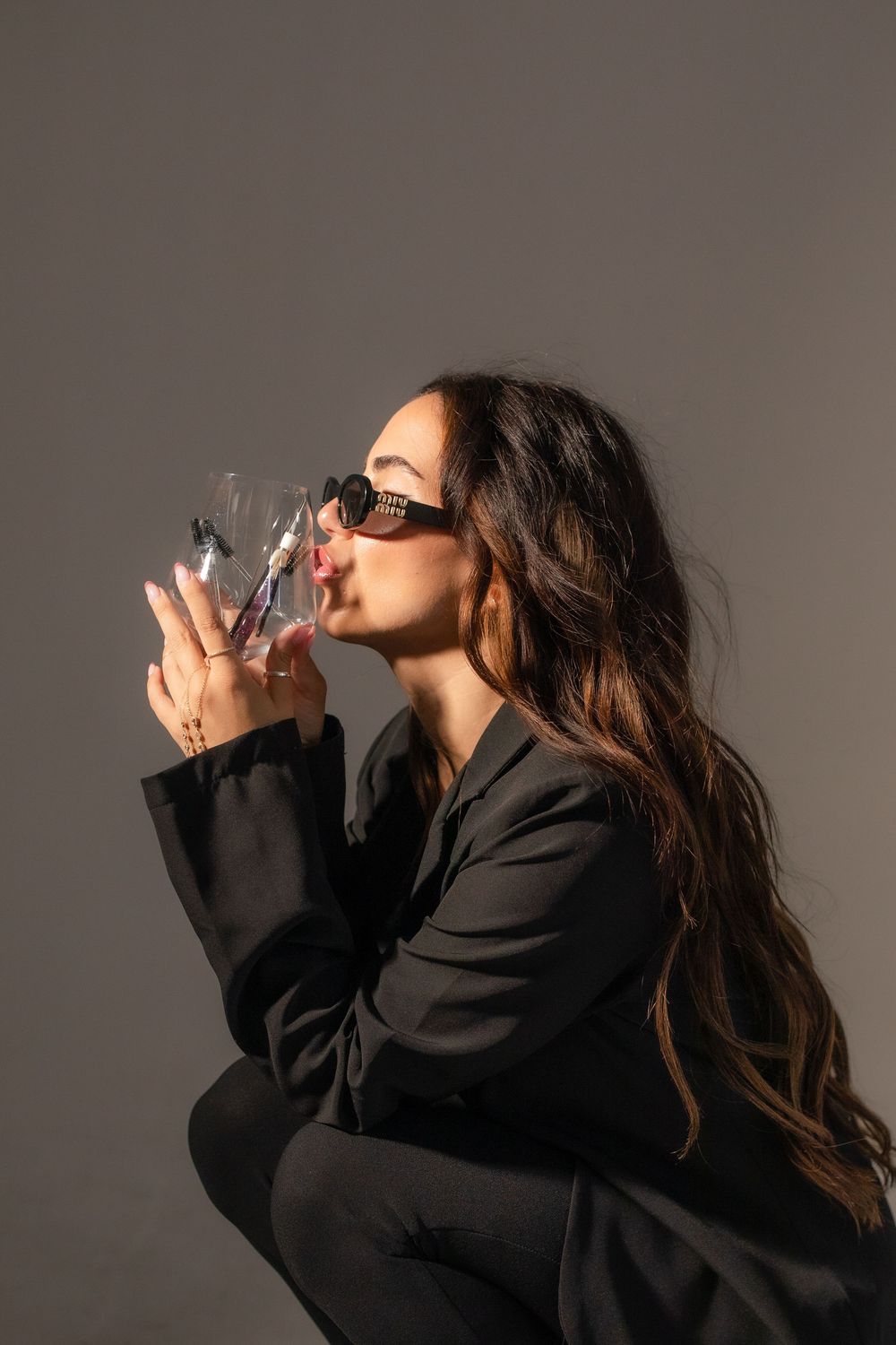 Stylish woman in black suit and sunglasses drinking from a glass, posed against a gray background.