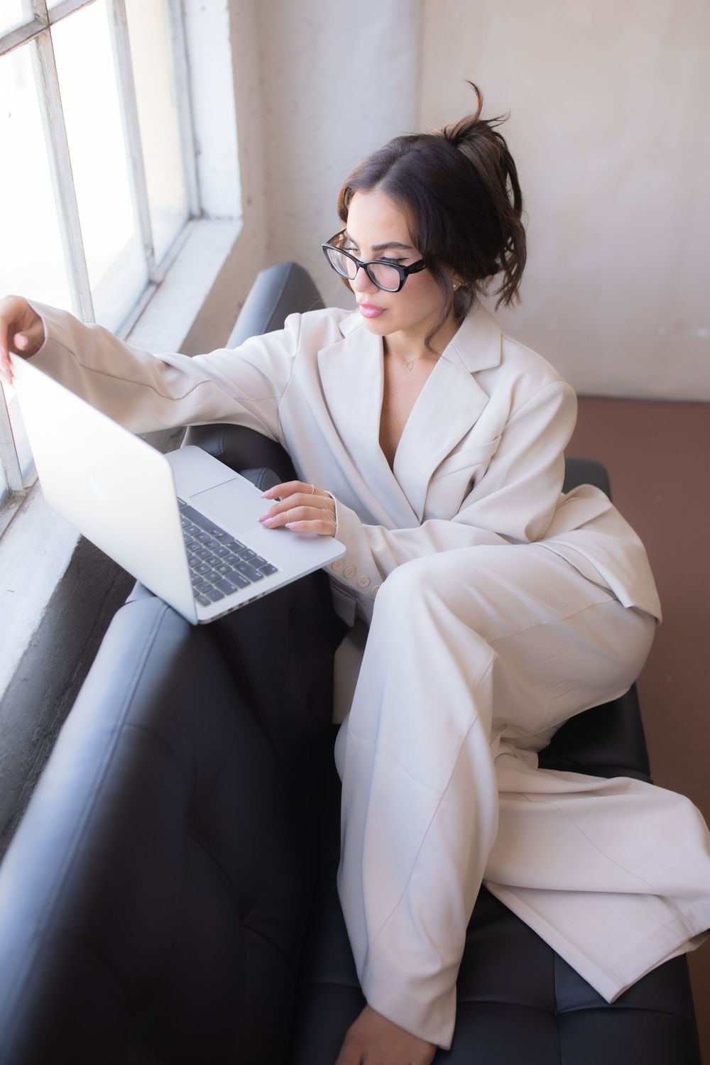 Professional woman in white suit working on laptop while seated on black sofa near window wearing glasses.