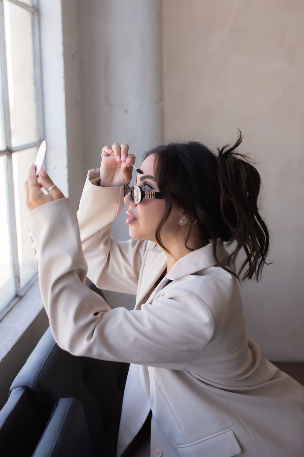 Stylish brunette woman in beige blazer and sunglasses taking a selfie near a window indoors.