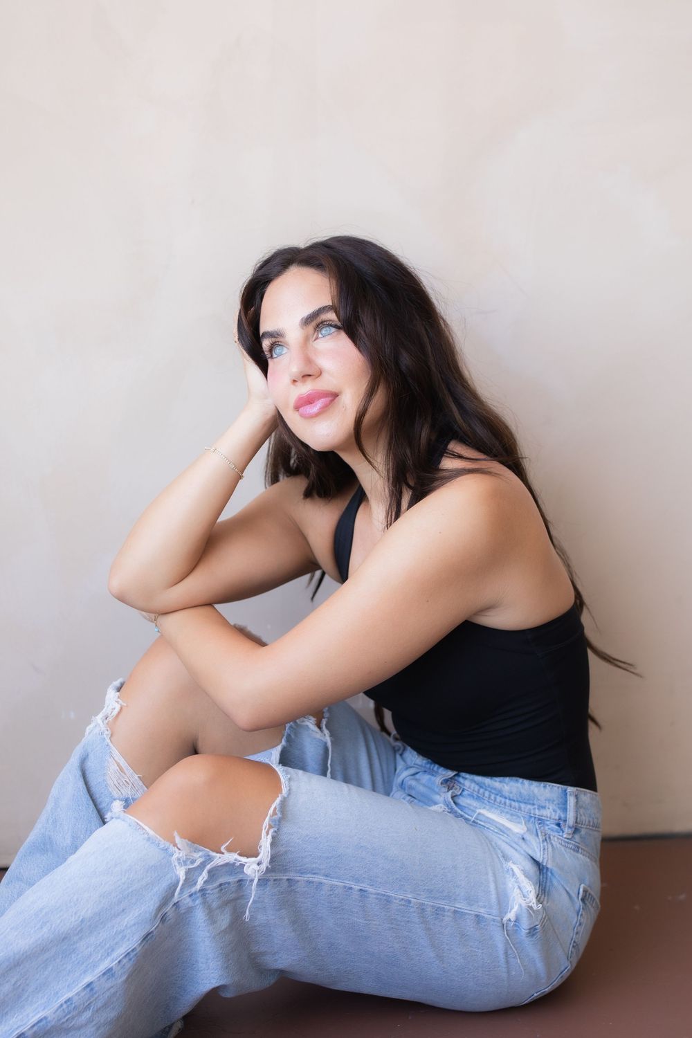Young woman with dark hair sitting against a wall in ripped jeans and black tank top, looking up thoughtfully.