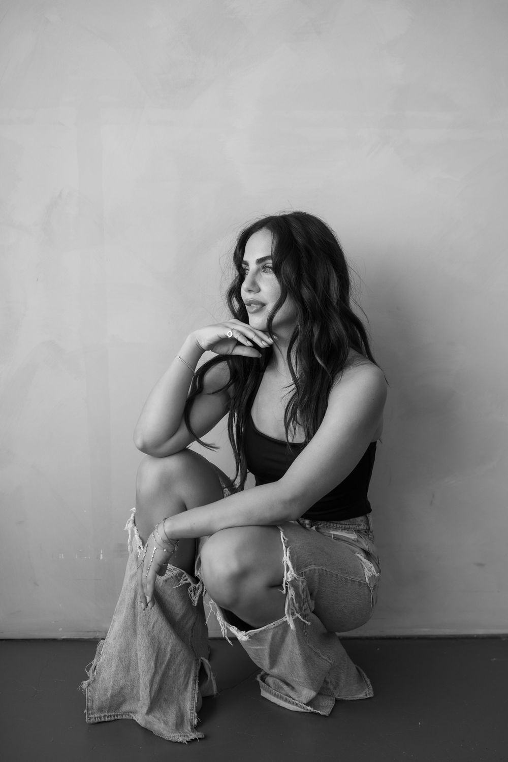 Black and white photo of a young woman in a black top and distressed flare jeans, sitting and gazing thoughtfully to the side.
