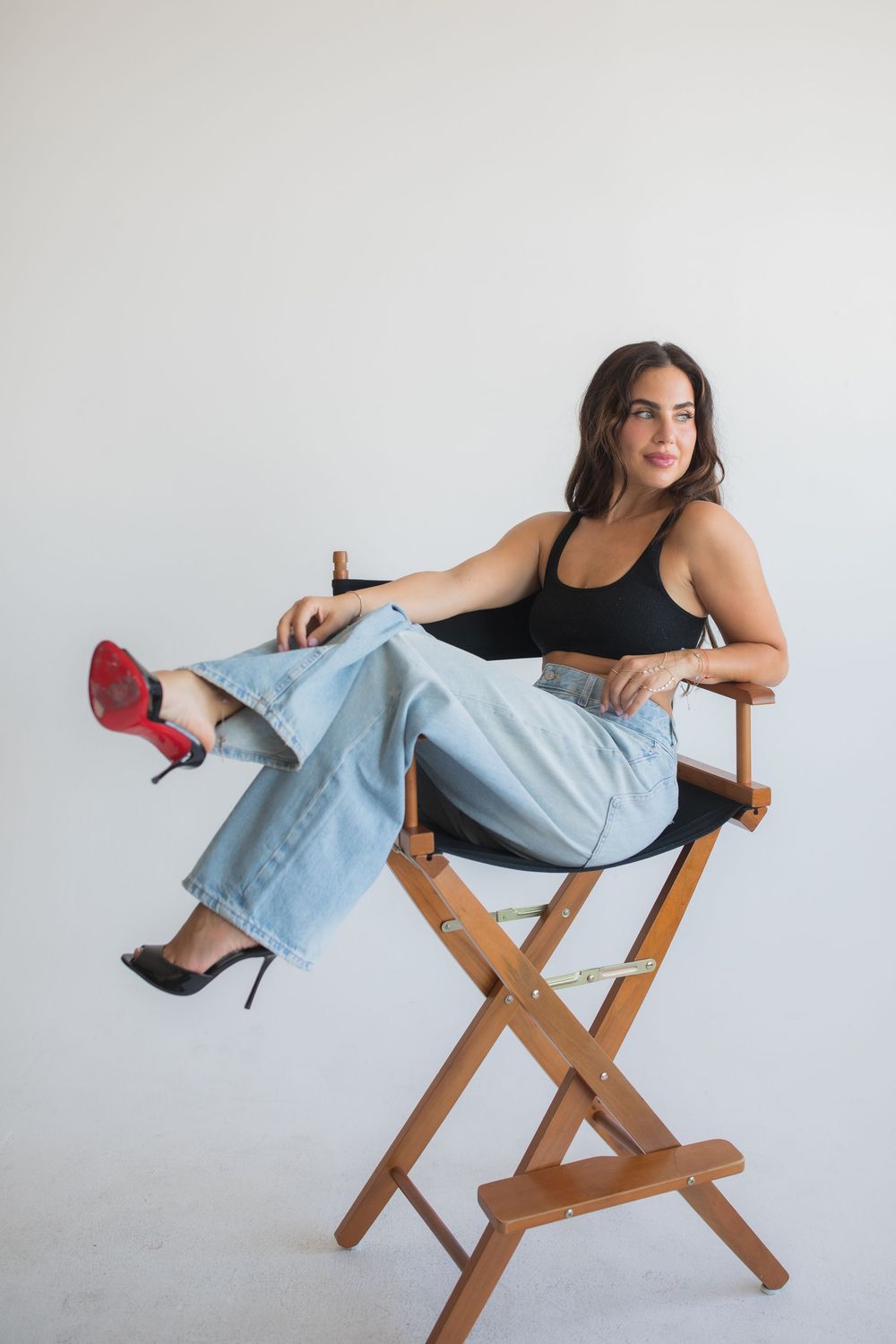 Woman in black crop top, wide-leg jeans, and mismatched heels sitting casually in a wooden director's chair.