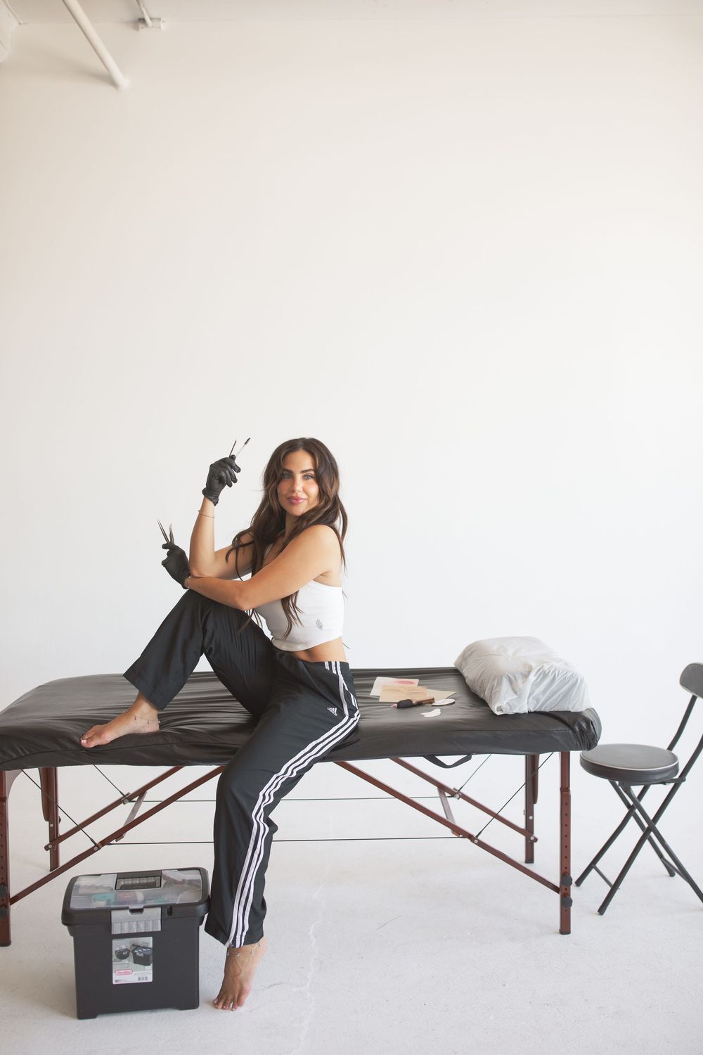 A young woman in a white crop top and black track pants sits on a massage table holding tattoo equipment in a bright studio.
