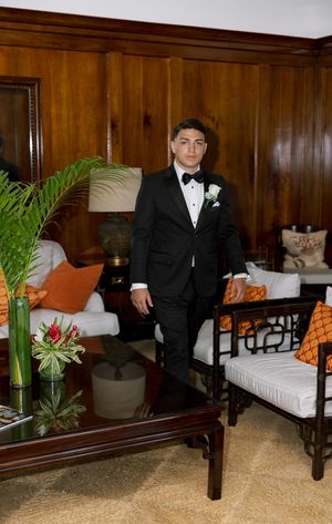Young man in black tuxedo with bow tie and boutonniere stands in elegant wood-paneled room with tropical decor.