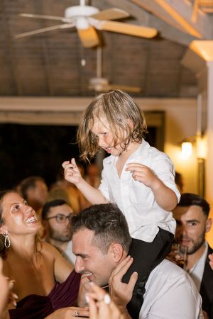 A young child in a white shirt dances joyfully on an adult's shoulders at a wedding reception.