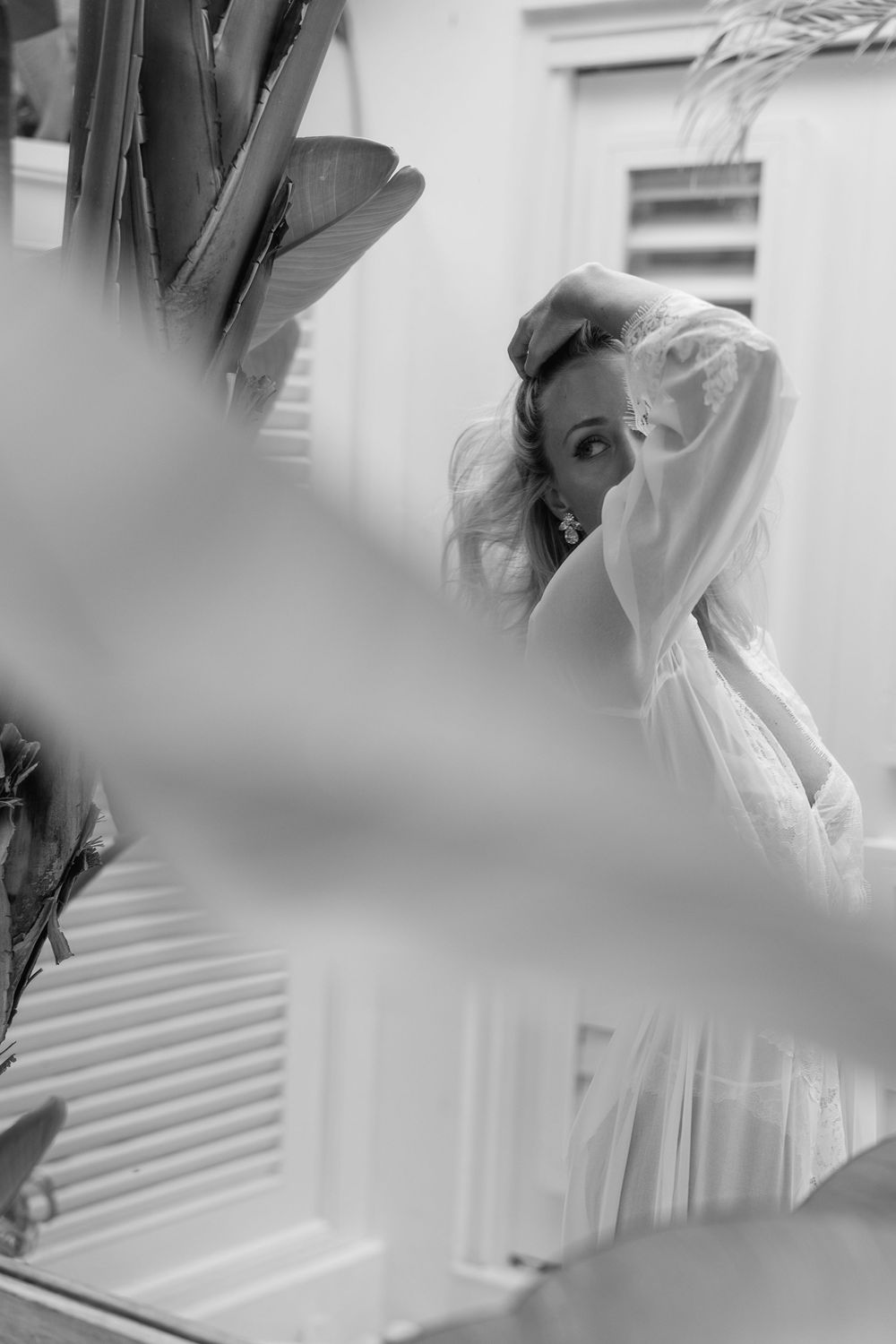 Black and white photo of a woman in a white lace dress posing near tropical plants and shuttered windows.