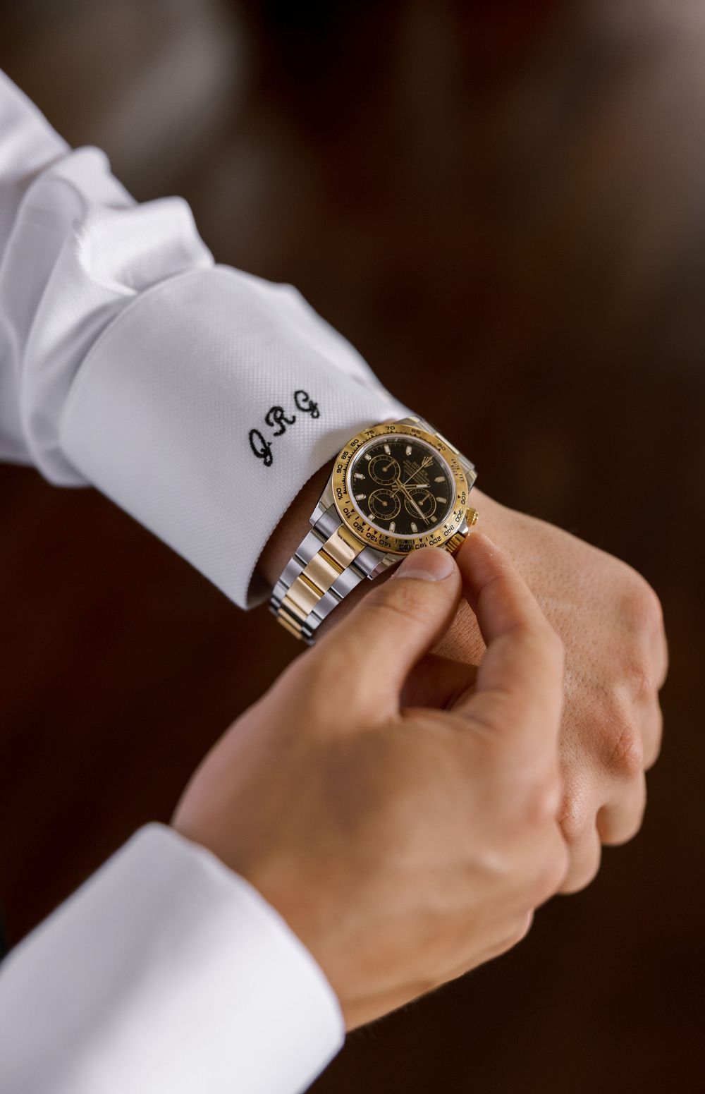 A man in a monogrammed white dress shirt adjusts a two-tone gold and silver Rolex Daytona watch on his wrist.