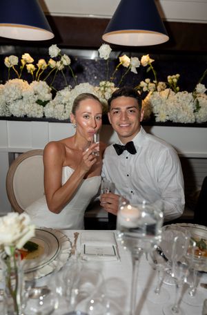 Bride and groom smiling at wedding reception table with champagne glasses, white floral backdrop, and elegant table settings.