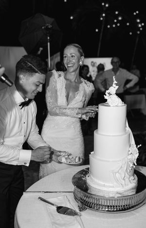 Bride and groom cutting elegant three-tier white wedding cake at outdoor reception in black and white photo.