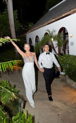 Couple in formal attire holding hands outdoors at night, woman in white jumpsuit raises arm joyfully near tropical venue.