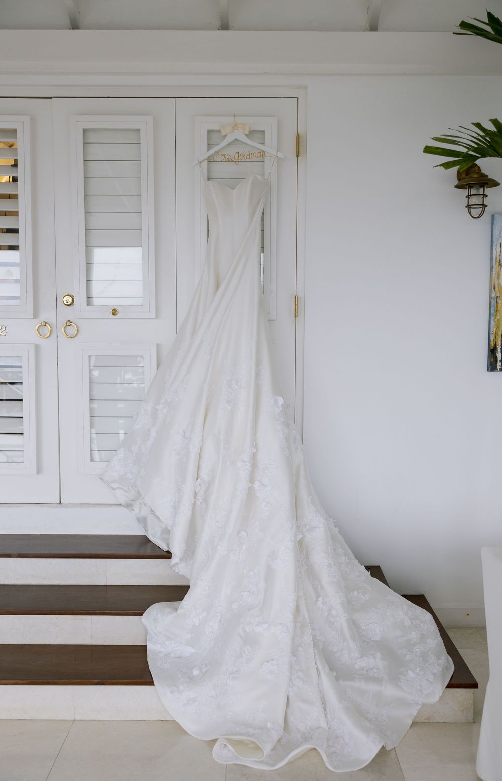 White lace wedding dress with long train hanging on decorative hanger against white shuttered doors in bright room.