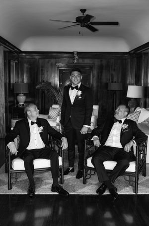 Black and white wedding photo of groom standing between two seated men in tuxedos in an elegant wood-paneled room.