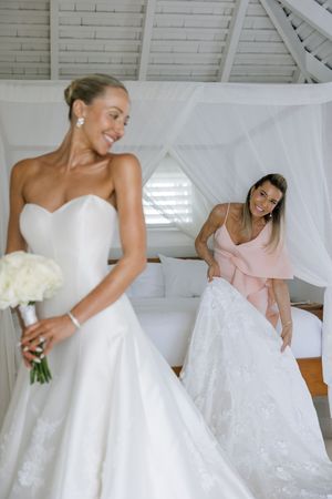 Bride in white strapless gown holds bouquet while bridesmaid in pink adjusts the train in a bright room.