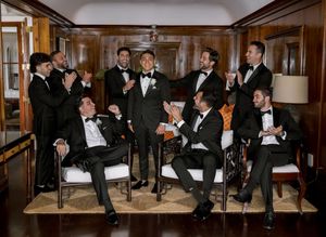 Groom and groomsmen in black tuxedos celebrate together in an elegant wood-paneled room at a wedding.