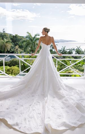 Bride in elegant white satin A-line wedding gown with long lace train stands on tropical resort balcony overlooking ocean.